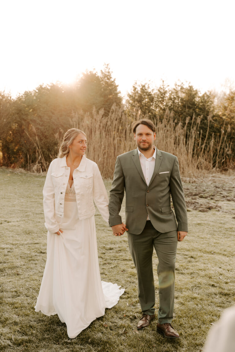 Braut in weißem Kleid und Jacke hält sich an den Händen mit Bräutigam in grünem Anzug, spazieren im Freien bei Sonnenuntergang in Brandenburg, umgeben von schöner Hochzeitsdeko.