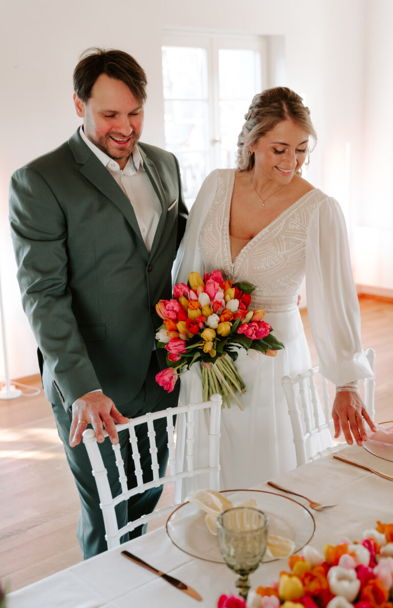 Eine Braut mit einem bunten Blumenstrauß steht lächelnd neben dem Bräutigam an einem wunderschön dekorierten Hochzeitstisch, der elegante Hochzeitsdeko von einem erfahrenen Hochzeitsplaner in Brandenburg zeigt.