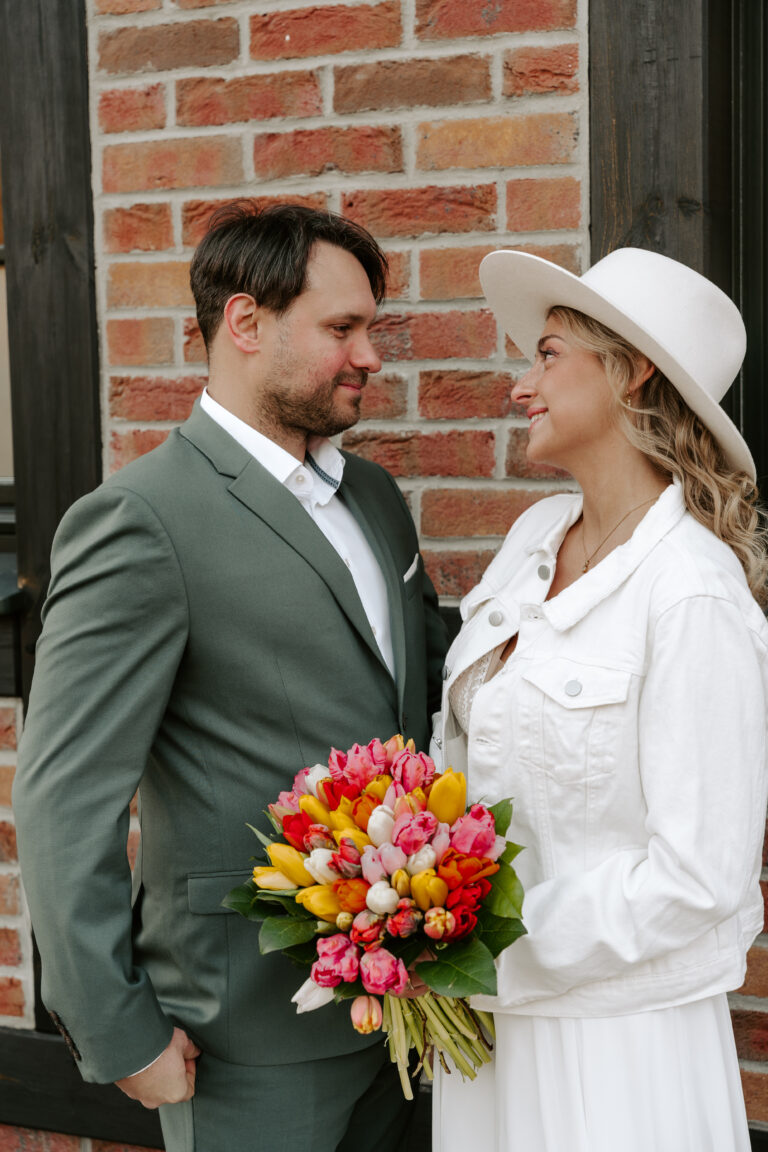 Ein Paar steht in Berlin lächelnd beieinander; sie hält einen Tulpenstrauß in der Hand und trägt einen weißen Hut und eine Jacke, um mit ihrer Freien Traurednerin zu feiern.