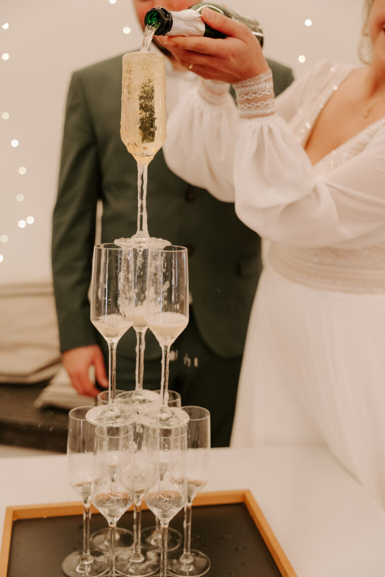 Eine Frau in Weiß schenkt Champagner in einen Turm von Gläsern ein, daneben steht ein Mann im Anzug - ein magischer Moment, den ein Hochzeitsplaner in Brandenburg eingefangen hat.