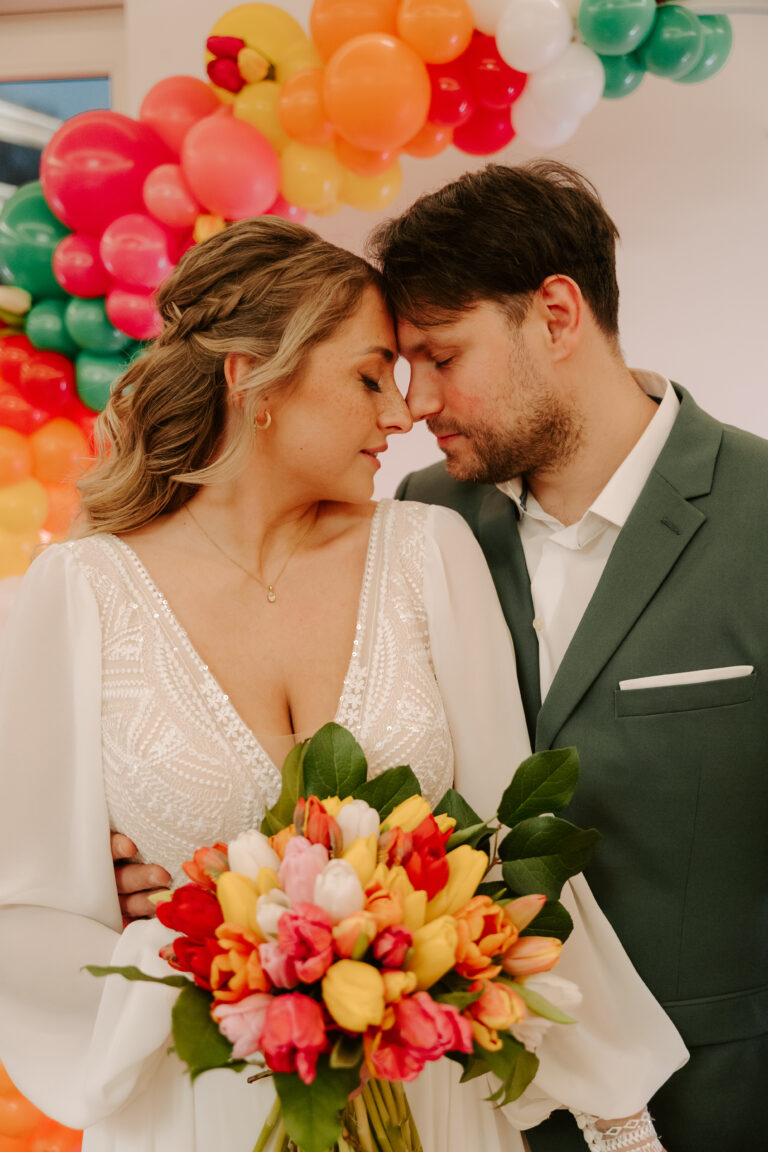 Ein Paar in Hochzeitskleidung umarmt sich mit einem farbenfrohen Blumenstrauß in der Hand, umgeben von bunten Luftballons und eleganter Hochzeitsdeko - und fängt damit die Freude an einer brandenburgischen oder Berliner Feier ein.