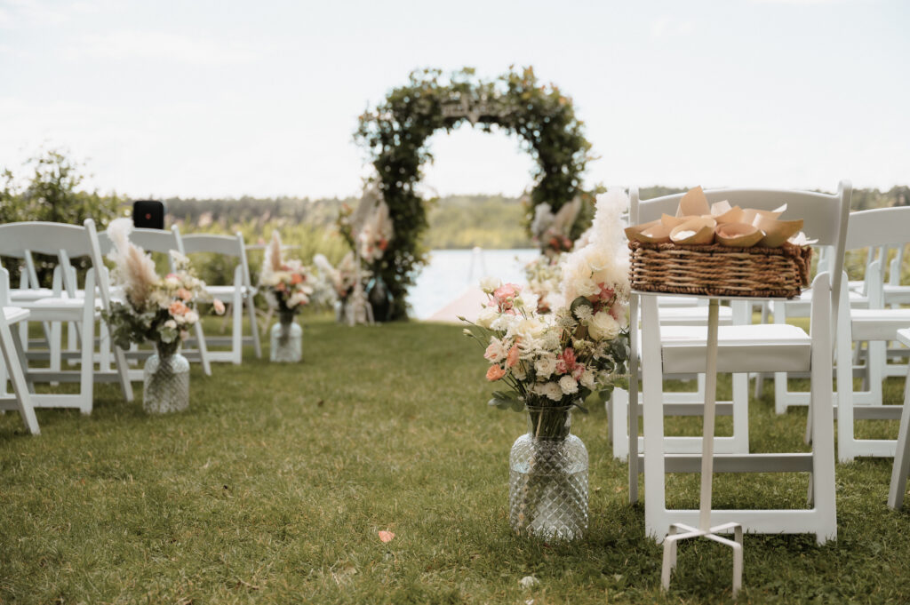 Hochzeit im Freien mit weißen Stühlen, Blumenarrangements und einem begrünten Bogen am Wasser - perfekt für eine romantische Zeremonie in Brandenburg, fachmännisch gestaltet von Ihrem Hochzeitsplaner.