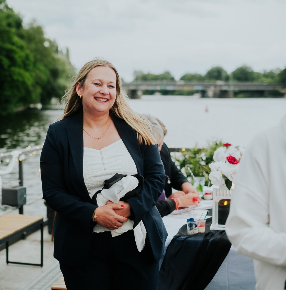 Frau im dunklen Anzug lächelnd an einem Fluss in Berlin, mit eleganter Hochzeitsdeko und Menschen an einem Tisch mit Blumen im Hintergrund.