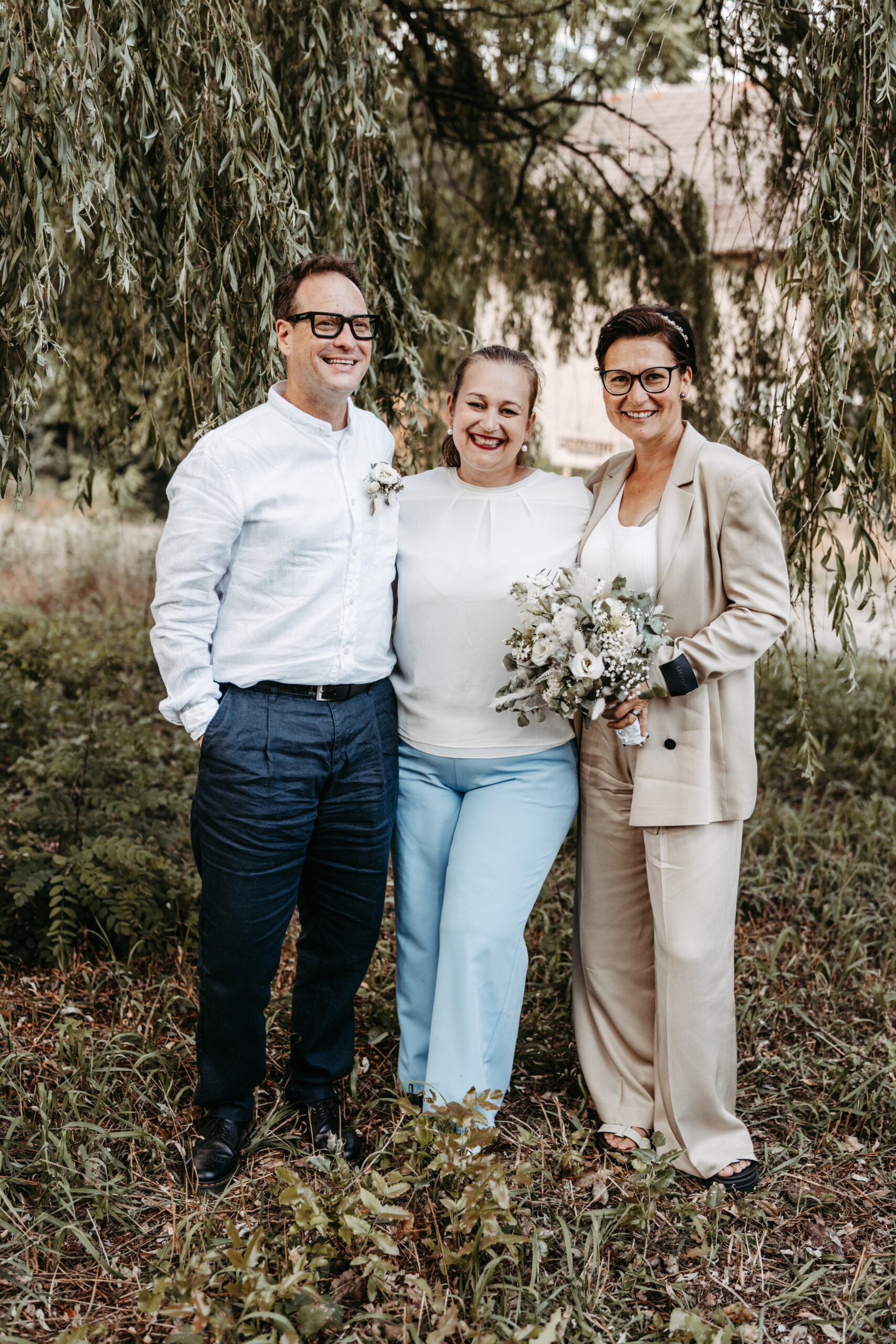 Drei lächelnde Erwachsene stehen im Freien in Brandenburg; einer hält einen Blumenstrauß, alle sind in leichte, halb-formelle Kleidung gekleidet - perfekt für einen Hochzeitsplaner oder eine Inspiration für eine Freie Traurednerin.