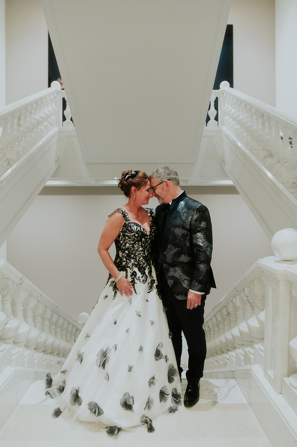 Ein formell gekleidetes Paar steht eng beieinander auf einer großen weißen Treppe in Brandenburg und lächelt sich liebevoll an, umgeben von eleganter Hochzeitsdeko.