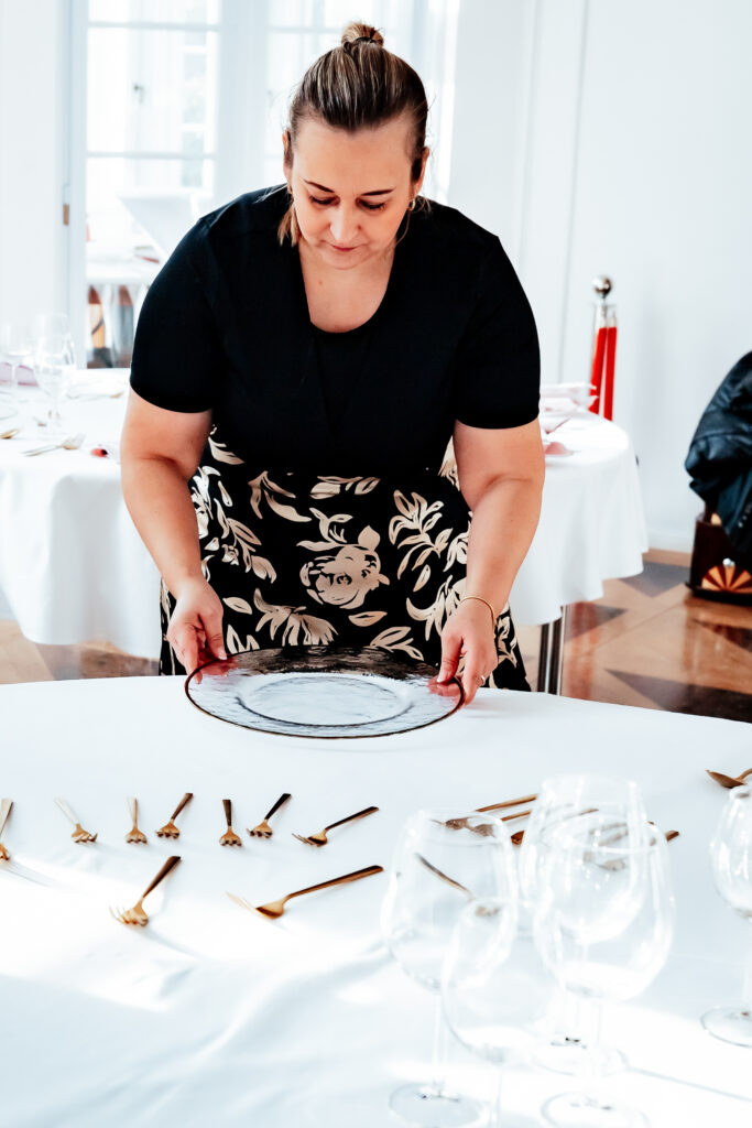 Eine Frau stellt einen durchsichtigen Teller auf einen mit Besteck und Weingläsern gedeckten Tisch und fügt so eine elegante Hochzeitsdeko in einem hellen Raum hinzu - perfekt für jeden Berliner Hochzeitsplaner, der Inspiration sucht.