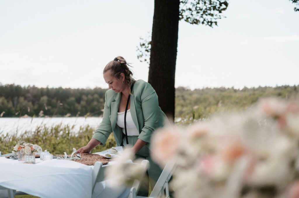 Eine Frau in einem grünen Anzug, vielleicht eine Hochzeitsplanerin, deckt einen Tisch im Freien in der Nähe eines Sees in Brandenburg, wobei Blumen und Bäume eine malerische Kulisse bilden.