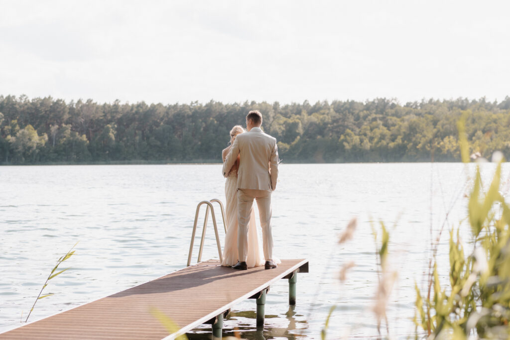 Ein leicht bekleidetes Paar steht auf einem Steg mit Blick auf einen ruhigen See in Brandenburg, mit Bäumen im Hintergrund.