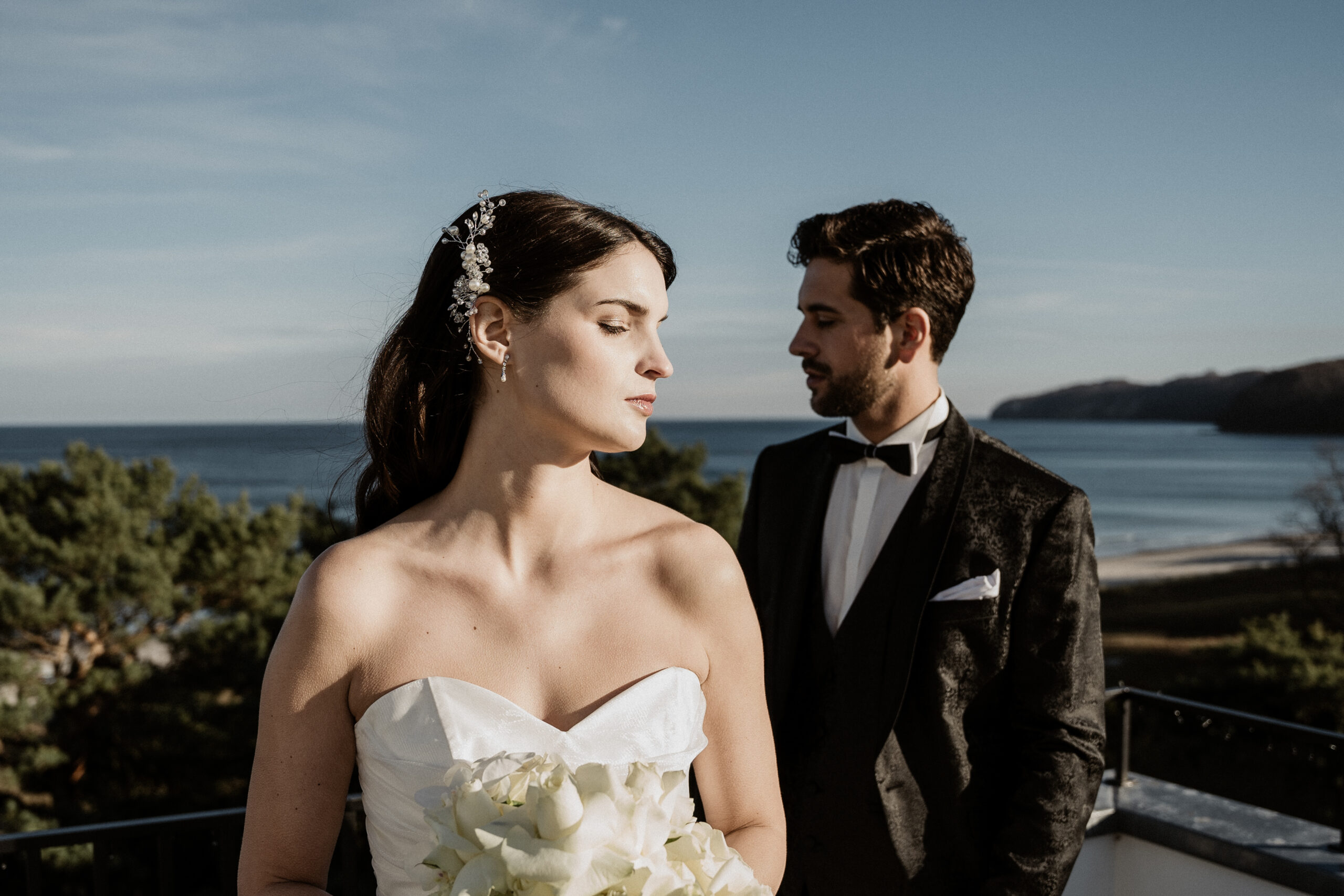 Die Braut im weißen Kleid und mit Blumen in der Hand steht vor dem Bräutigam auf dem Balkon mit Blick auf das Meer und die Küste. Die elegante Hochzeitsdeko ist vom modernen Stil Berlins inspiriert.