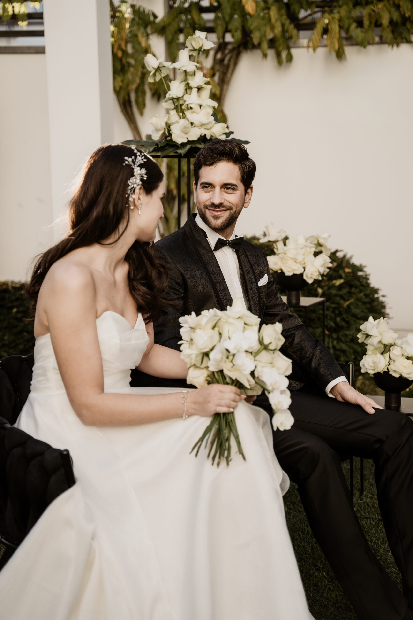 Braut und Bräutigam sitzen lächelnd beieinander und halten weiße Rosen in der Hand, umgeben von eleganter Hochzeitsdeko und prächtigen weißen Blumenarrangements in Berlin.