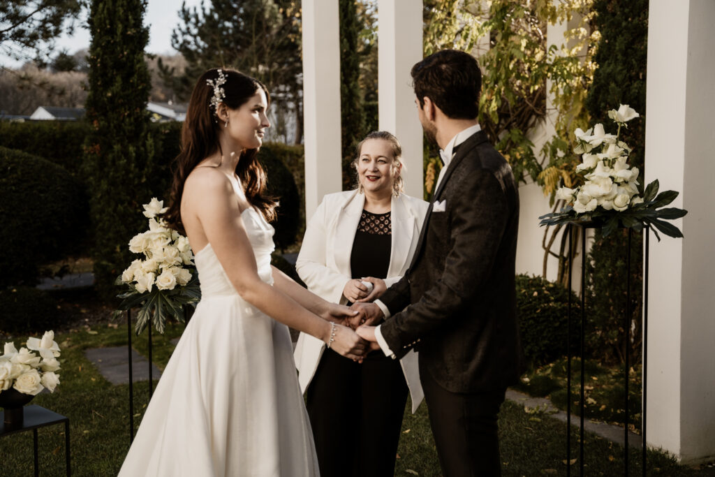 Eine Braut und ein Bräutigam halten sich an den Händen und lächeln sich während einer Hochzeitszeremonie im Freien in Brandenburg an, umgeben von einer wunderschönen Hochzeitsdeko.