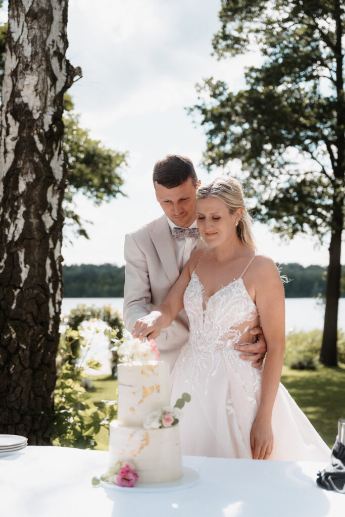 Eine Braut und ein Bräutigam schneiden ihre Hochzeitstorte an einem sonnigen Tag im Freien an einem See an, umgeben von einer wunderschönen Hochzeitsdeko, die von ihrem Hochzeitsplaner arrangiert wurde.