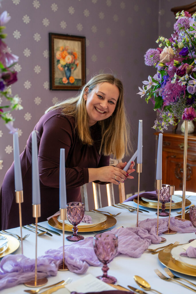Eine Frau lächelt, während sie einen lilafarbenen, eleganten Tisch mit Blumen und Kerzen arrangiert und damit ihr Talent als Hochzeitsplanerin für eine schöne Hochzeitsdeko unter Beweis stellt.