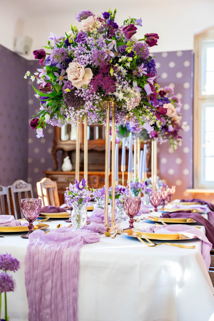 Elegant gedeckter Tisch mit hohen lilafarbenen Blumensträußen, fliederfarbenem Tischläufer und alten Gläsern - perfekt für eine romantische Berliner Feier, die von einer Freien Traurednerin geleitet wird.