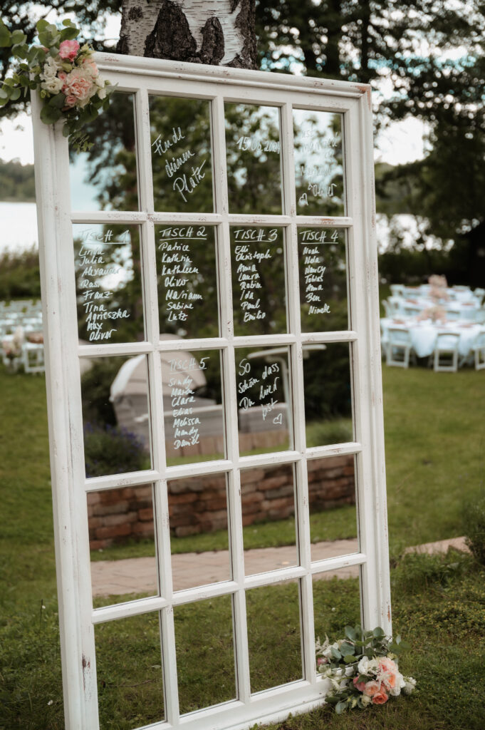 Eine alte Fensterscheibe, auf der die Sitzordnung für die Hochzeit aufgeschrieben ist, wunderschön mit Blumen dekoriert - perfekt für Paare in Brandenburg oder unter Anleitung eines Hochzeitsplaners oder einer Freien Traurednerin.