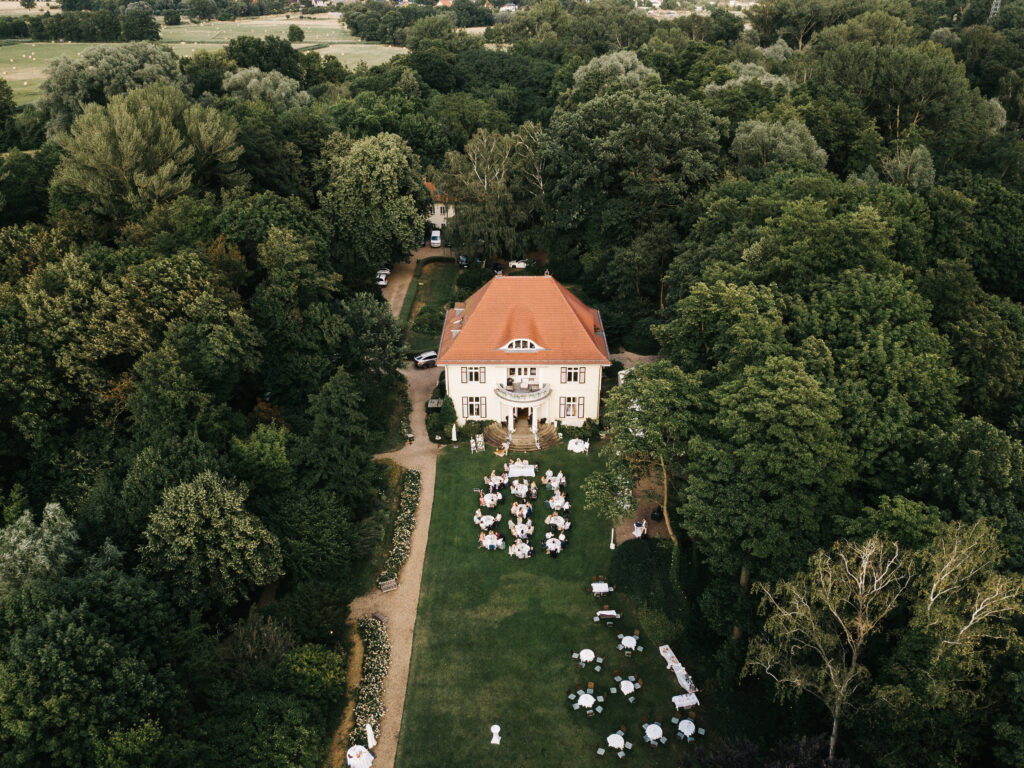 Luftaufnahme einer Gartenparty mit weißen Tischen vor einem großen Haus in Berlin, umgeben von dichten grünen Bäumen - die perfekte Inspiration für jeden Hochzeitsplaner oder jede Freie Traurednerin.