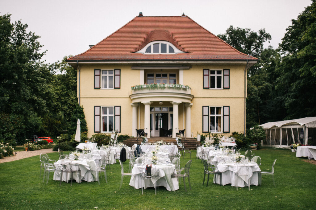 Elegante Hochzeitsfeier im Freien mit runden Tischen vor einem großen gelben Haus mit rotem Dach.