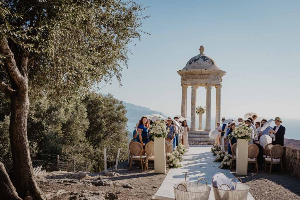 Hochzeitszeremonie im Freien mit Gästen, die an einem Blumengang sitzen, vor einem malerischen, küstennahen Steinpavillon - perfekt für Ihren mediterranen Hochzeitstraum und eine unvergessliche Freie Trauung Mallorca.
