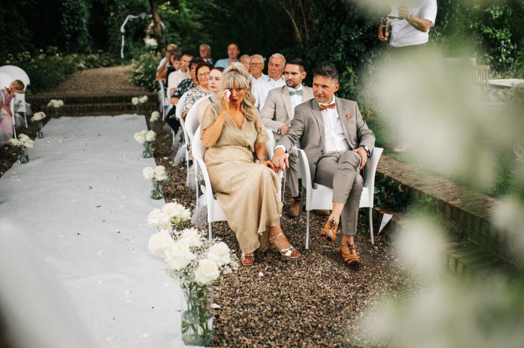 Die Gäste sitzen bei einer Hochzeitszeremonie im Freien in Reihen; eine Frau im Vordergrund wischt sich die Augen und sitzt neben einem Mann.