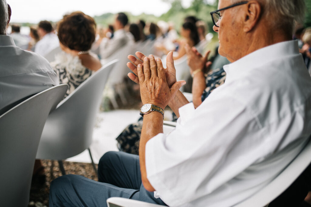 Älterer Mann mit Brille und weißem Hemd klatscht bei einer Veranstaltung im Freien mit anderen sitzenden Menschen.