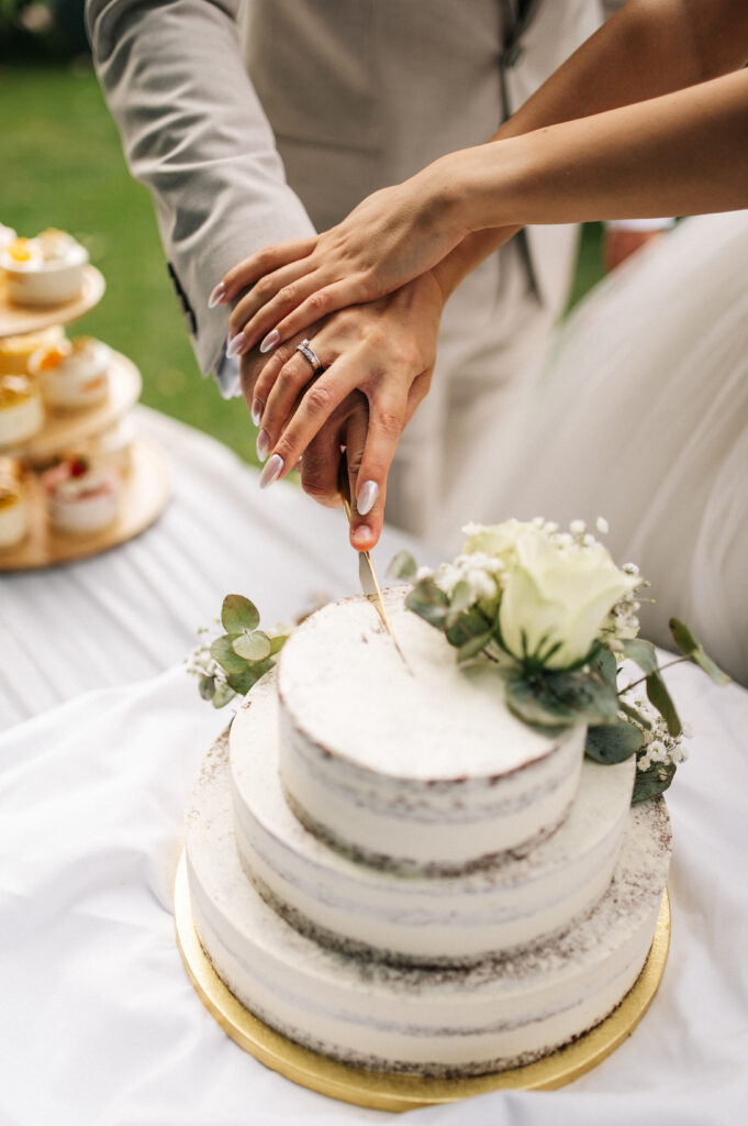 Ein Paar schneidet gemeinsam eine weiße Hochzeitstorte mit einem Messer an, wobei sich die Hände über der Torte überlappen.
