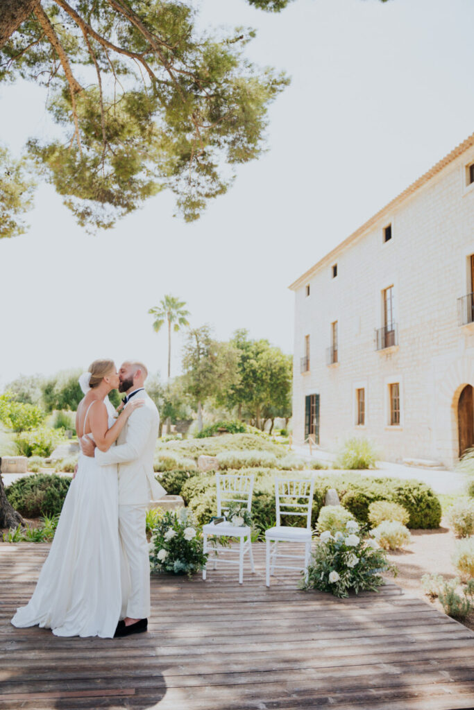 Braut und Bräutigam in weißer Kleidung umarmen sich im Freien auf einer Holzterrasse, umgeben von Grün und einem Steingebäude, das einen mediterranen Hochzeitstraum schafft - perfekt für eine Freie Trauung auf Mallorca.