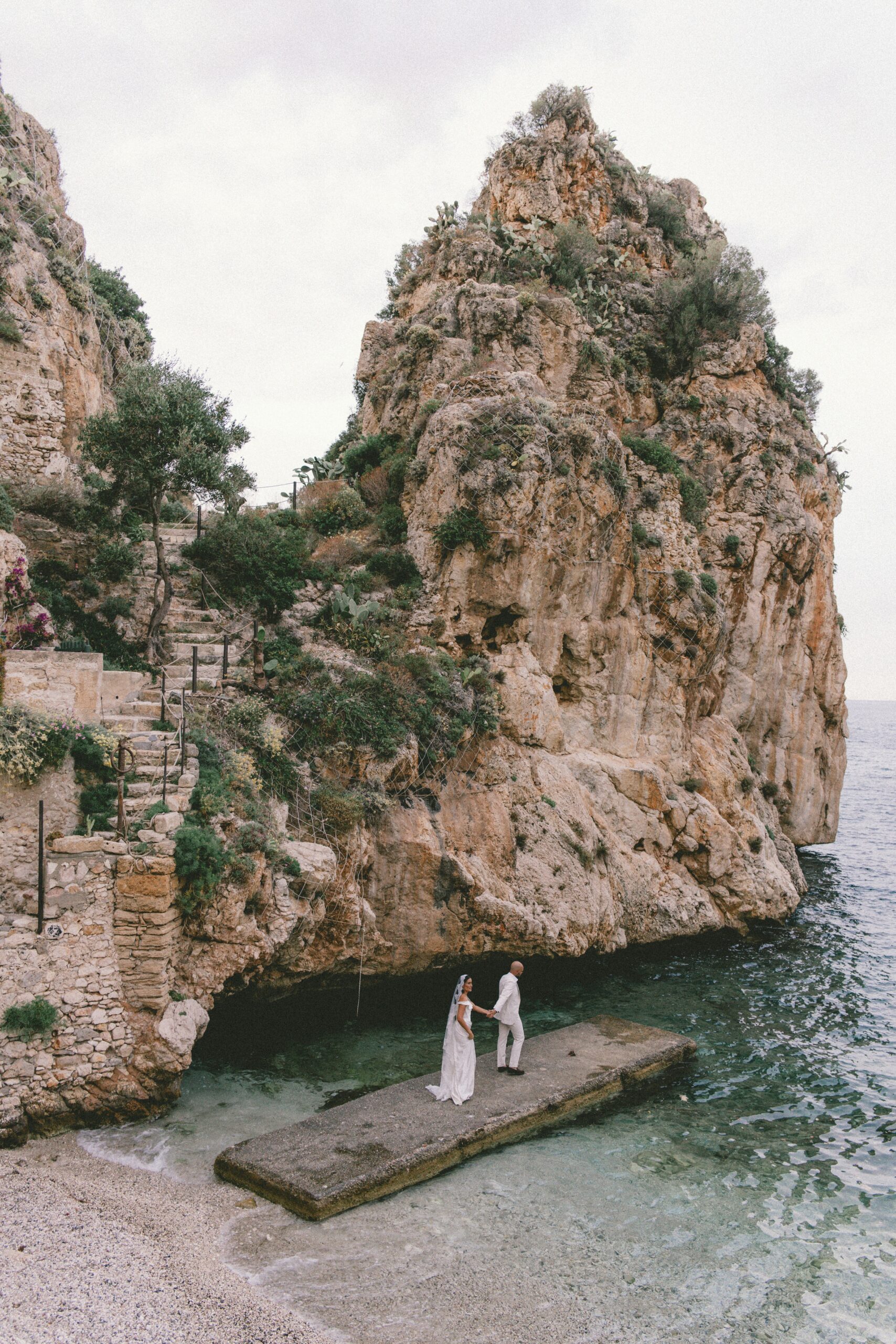 Ein Paar steht auf einem steinernen Steg am Meer, umrahmt von felsigen Klippen und üppigem Grün - ein wahrer mediterraner Hochzeitstraum und der perfekte Ort für eine Freie Trauung Mallorca.