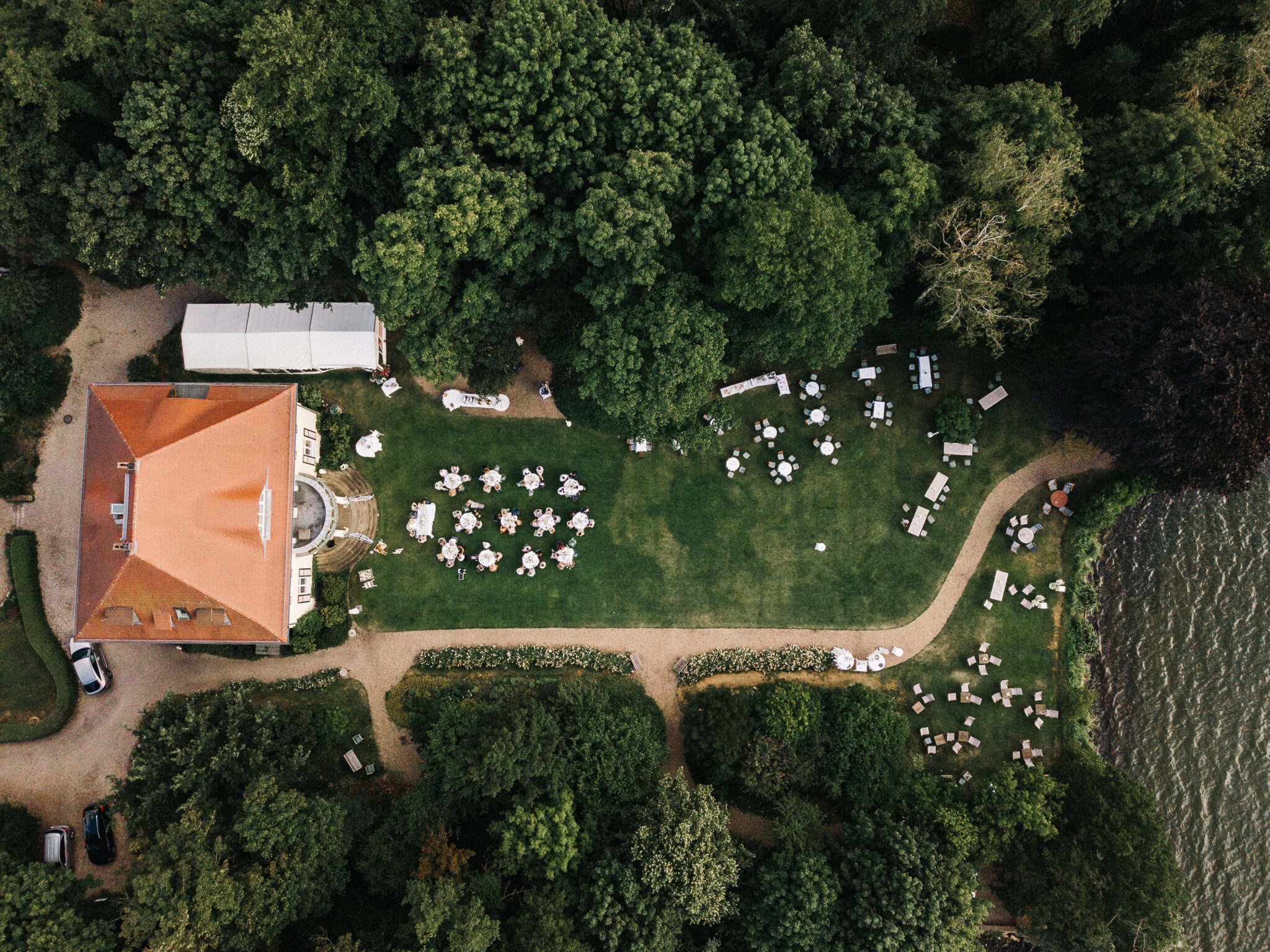 Luftaufnahme einer Gartenveranstaltung mit Tischen, die auf einer Wiese neben einem Gebäude aufgestellt und von Bäumen umgeben sind.