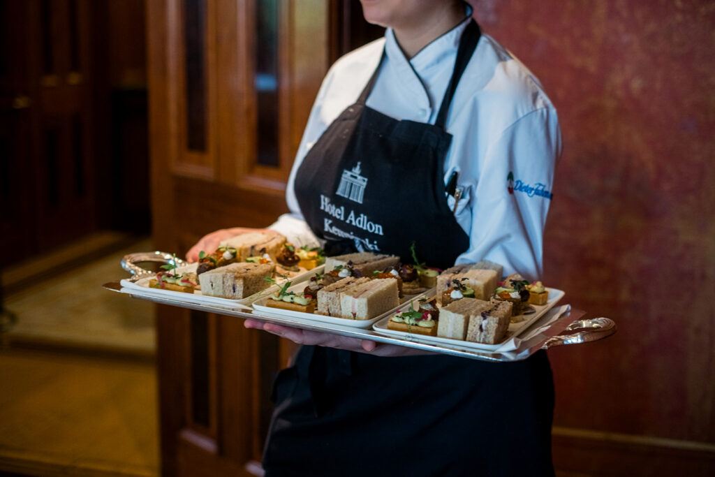 Ein Kellner in Uniform trägt im Hotel Adlon Berlin ein Tablett mit verschiedenen Sandwiches und Vorspeisen in einem formellen Rahmen.
