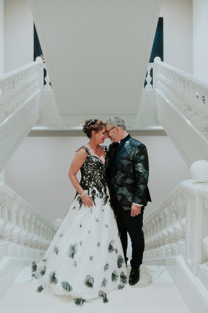Ein formell gekleidetes Paar steht eng beieinander auf einer weißen Treppe im Hotel Adlon Berlin, lächelt und berührt sich an der Stirn.