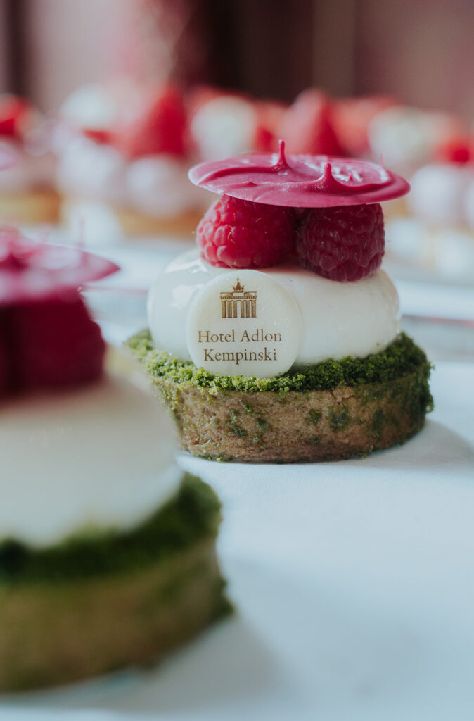 Miniteiggebäck mit weißer Sahne, frischen Himbeeren und einem Schokoladenmedaillon des Hotel Adlon Berlin darauf.