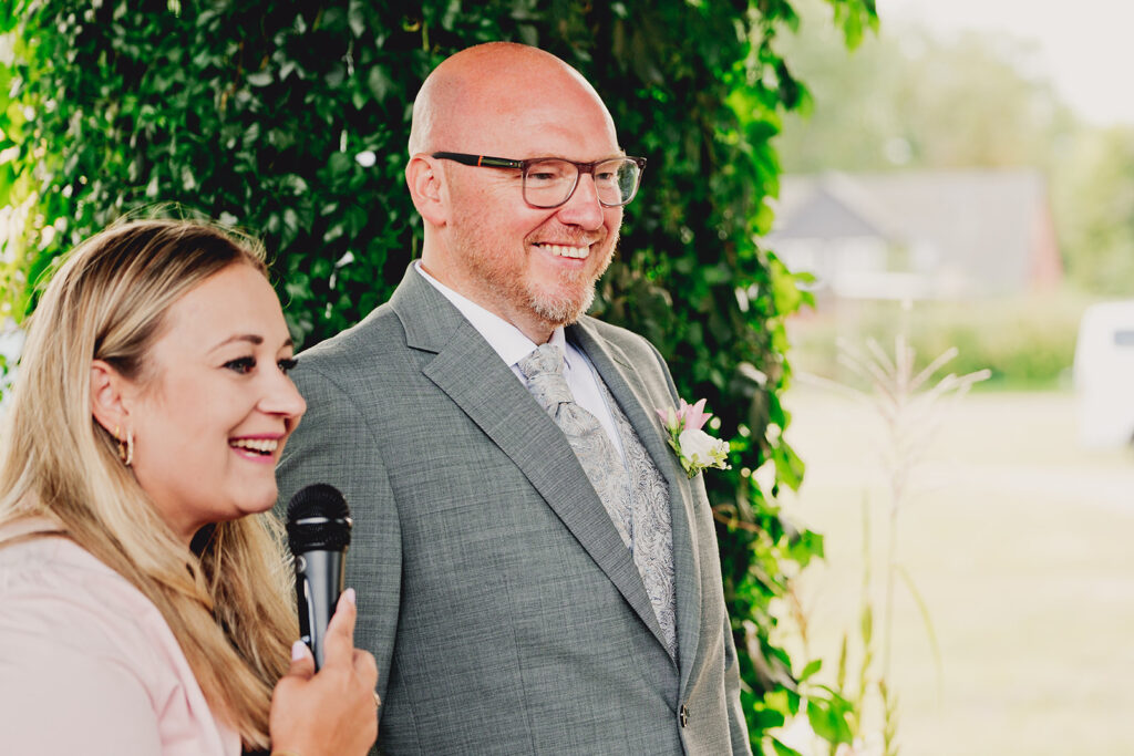 Eine lächelnde Frau mit einem Mikrofon steht neben einem Mann im Anzug mit einer Boutonniere während einer Freien Trauung in der Brennerei Genshagen, im Freien.