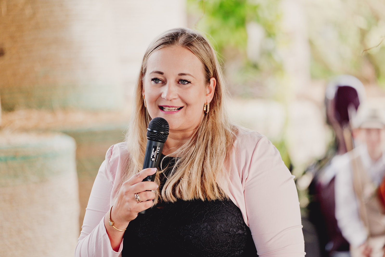 Eine Frau mit langen blonden Haaren hält ein Mikrofon und lächelt, während sie bei einer Veranstaltung der Freien Trauung in der Brennerei Genshagen spricht.