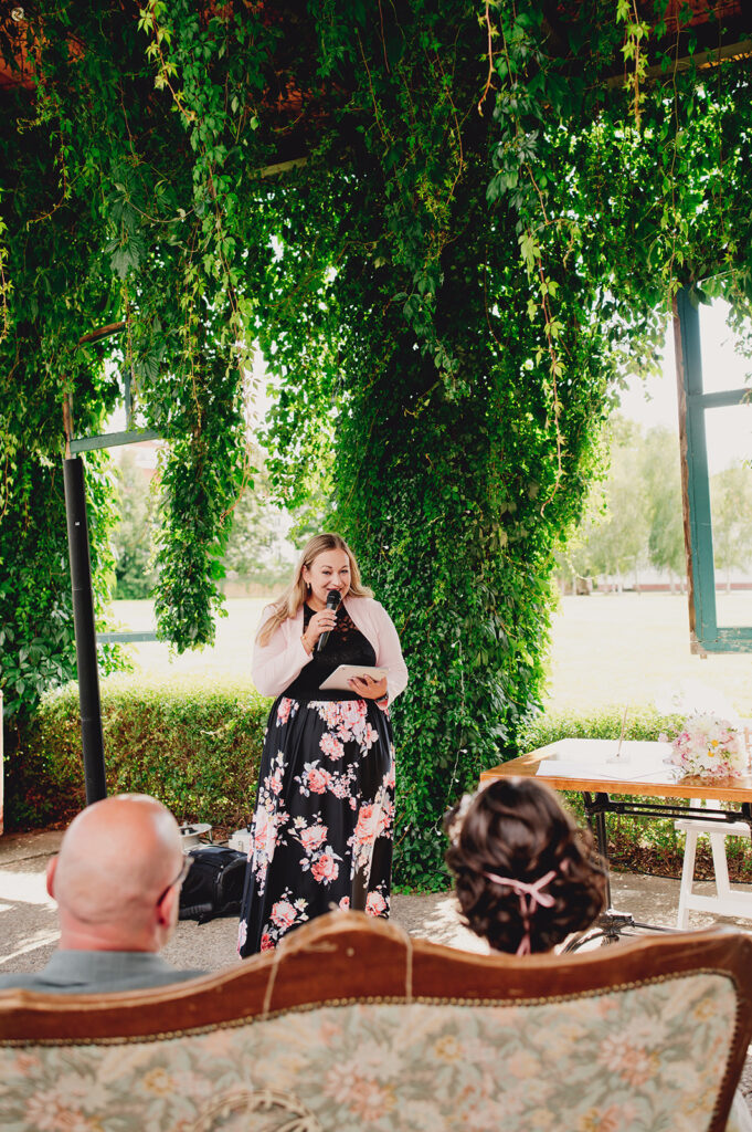 Eine Frau in einem geblümten Kleid spricht während einer Freien Trauung im Freien in ein Mikrofon. Im Vordergrund stehen Grünpflanzen und Gäste in der Brennerei Genshagen.