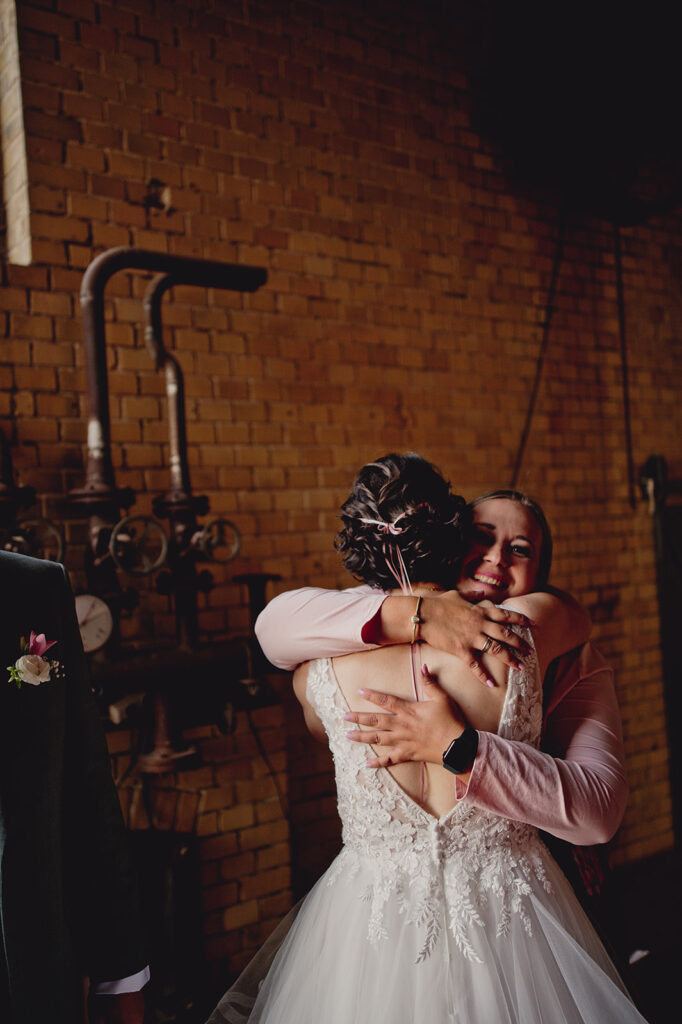 Zwei Frauen in formeller Kleidung umarmen sich nach einer herzlichen Freien Trauung vor einer Backsteinmauer mit Industrierohren in der Brennerei Genshagen.
