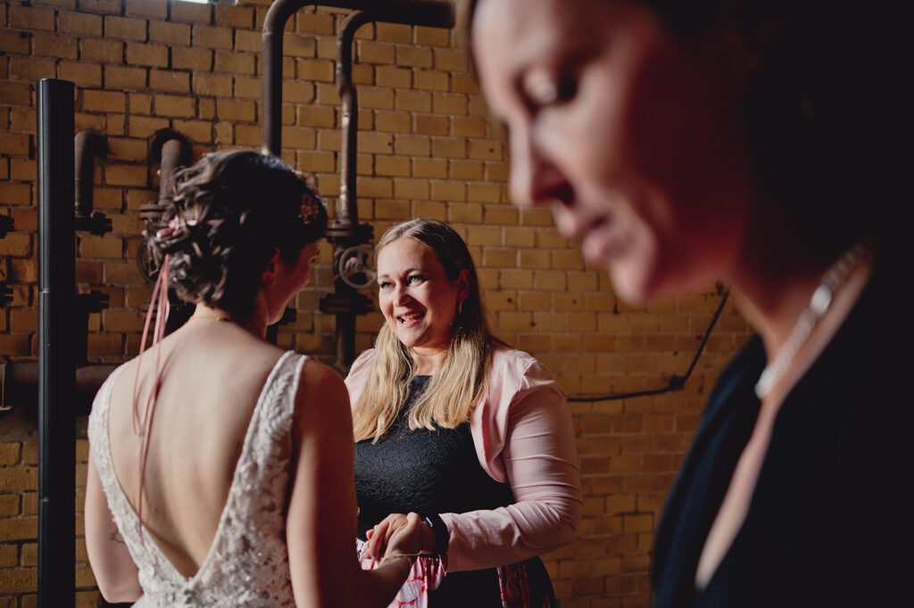 Eine Frau im Hochzeitskleid unterhält sich während einer Freien Trauung in der Brennerei Genshagen mit einem lächelnden Gast, während eine andere Frau unscharf im Vordergrund zu sehen ist.