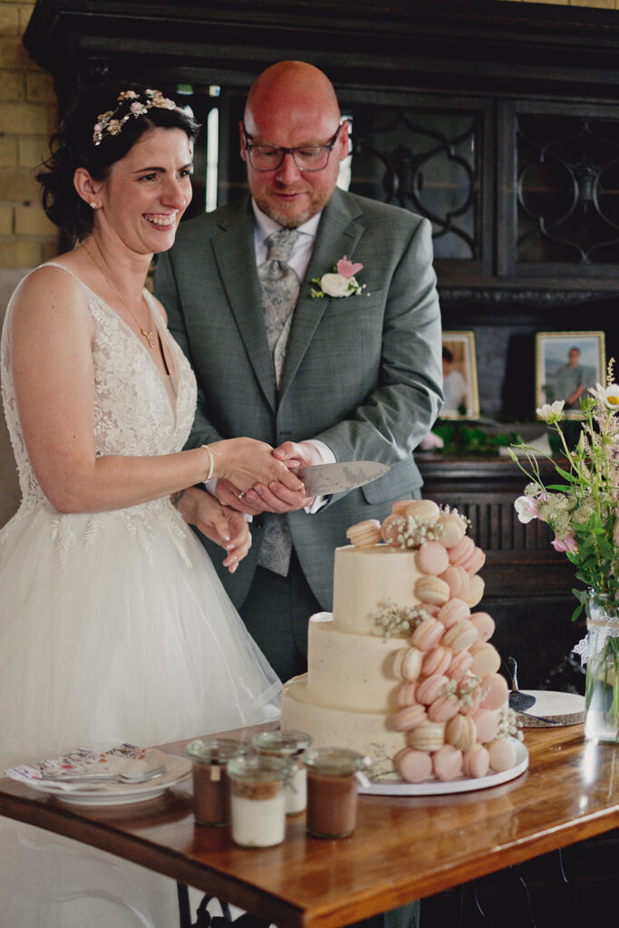 Braut und Bräutigam lächelnd beim Anschneiden einer mit Macarons und Blumen verzierten Hochzeitstorte während ihrer Freien Trauung in der Brennerei Genshagen.