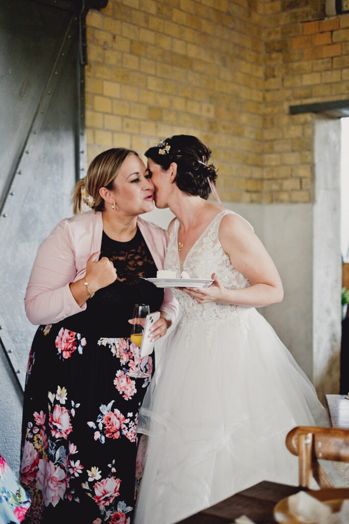 Eine Braut in einem weißen Kleid küsst eine Frau in einem geblümten Rock und einer rosafarbenen Jacke. Beide lächeln während ihrer Freien Trauung in der Brennerei Genshagen.