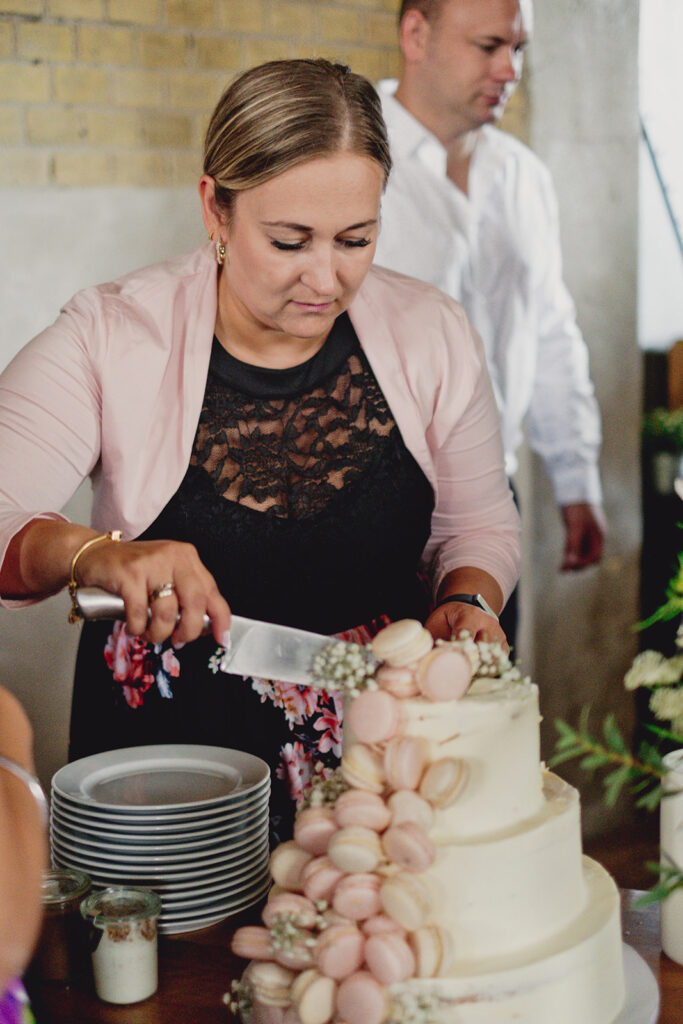 Bei einer Indoor-Veranstaltung schneidet eine Frau eine weiße Stufentorte an, die mit rosa Macarons und Blumen verziert ist, um eine Freie Trauung in der Brennerei Genshagen zu feiern.