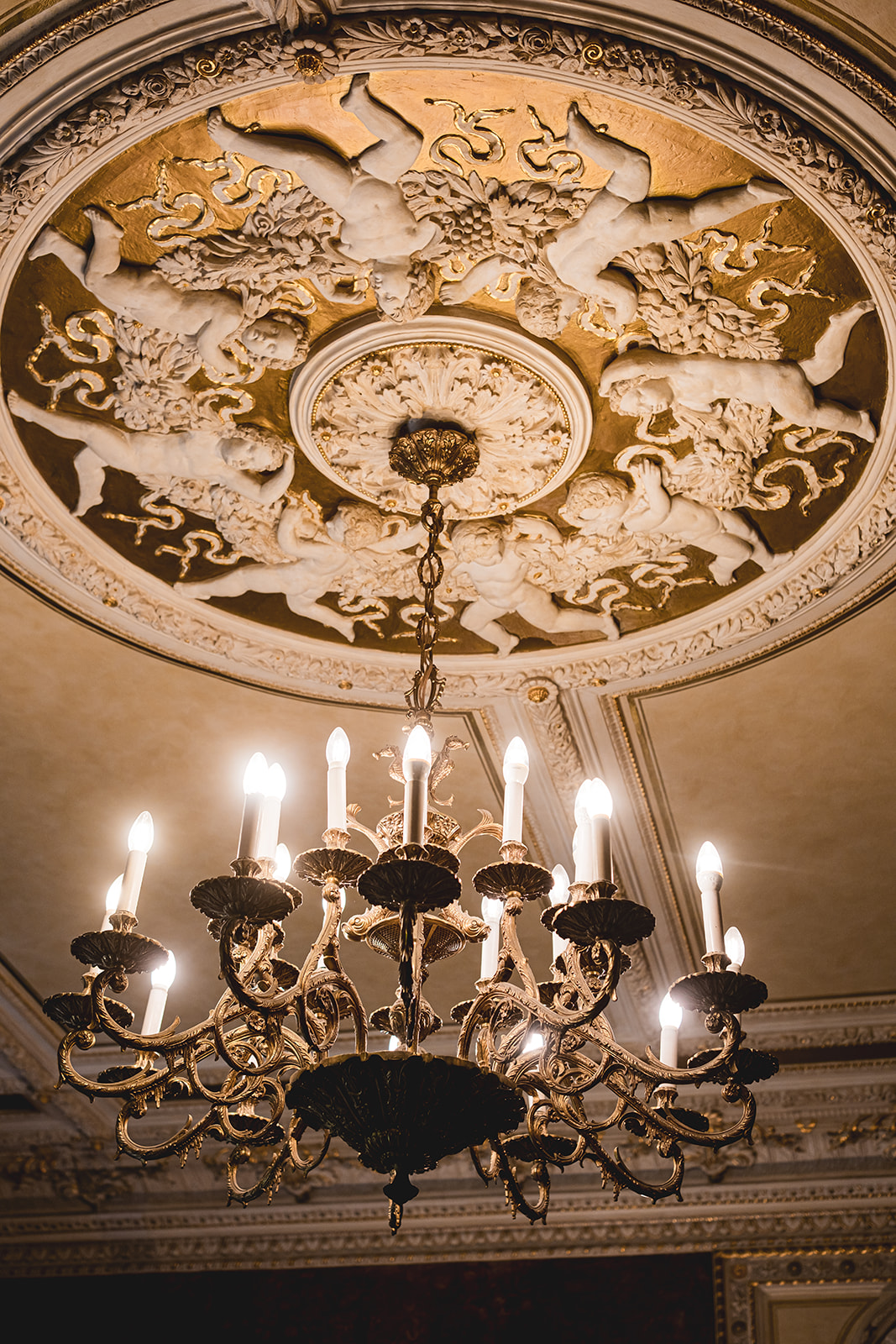 Verschnörkelter Kronleuchter mit brennenden Kerzen, die von einer dekorativen gold-weißen Decke mit geschnitzten Figuren herabhängen. Perfekt für eine Standesamt-Zeremonie oder Freie Trauung in einem eleganten Rahmen.