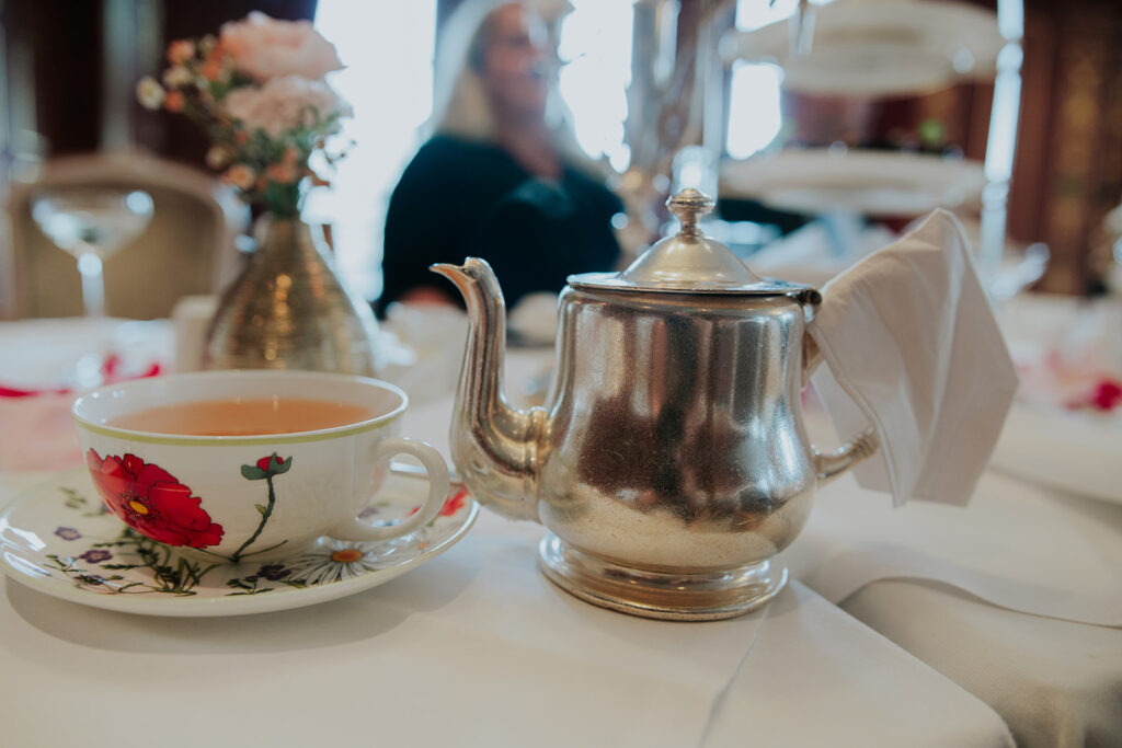 Eine silberne Teekanne und eine geblümte Teetasse stehen auf einem weißen Tischtuch mit Blumen im Hintergrund und erinnern an die zeitlose Eleganz des Hotel Adlon Berlin.