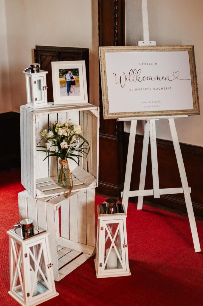 Ein Hochzeits-Willkommensschild, weiße Laternen, gerahmte Fotos und ein Blumenstrauß auf weißen Holzkisten - die perfekte Hochzeitsdeko für Ihre Hochzeit, auch erhältlich im Dekoverleih.