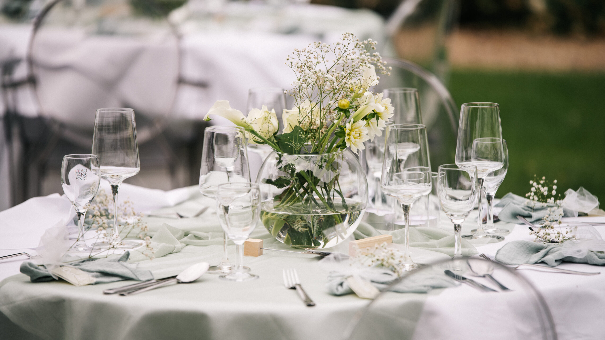 Elegant gedeckter Tisch im Freien mit Glaswaren, weißen Blumen in einer Vase und zartgrünen Servietten.