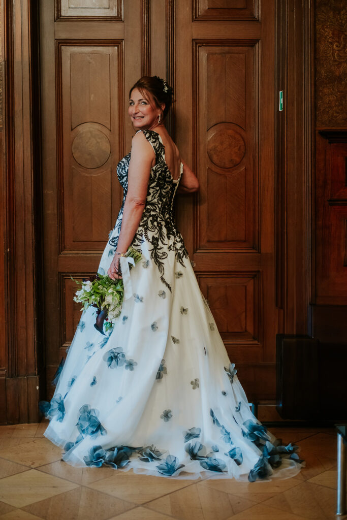 Frau in weißem Kleid mit schwarzen Blumendetails, einen Blumenstrauß in der Hand, vor Holztüren stehend - perfekt für eine Feier des Standesamtes oder der Freien Trauung.