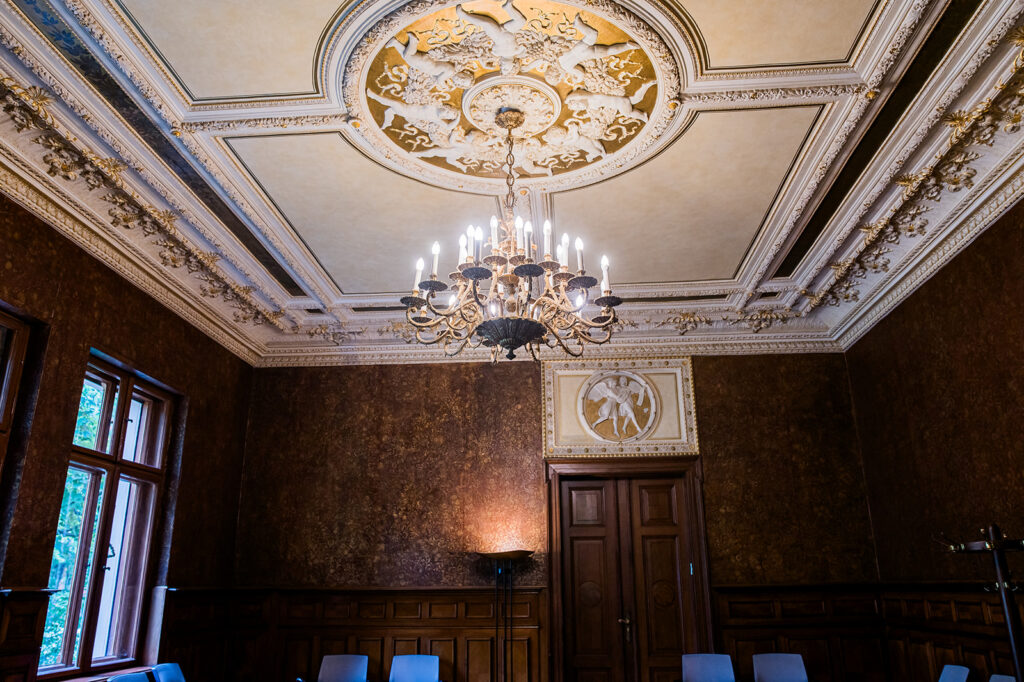 Eleganter Raum mit verzierter Decke, Kronleuchter, Holzvertäfelung und darunter angeordneten Stuhlreihen - ein idealer Rahmen für ein Standesamt oder eine Freie Trauung in der von Ihnen gewählten Trauform.