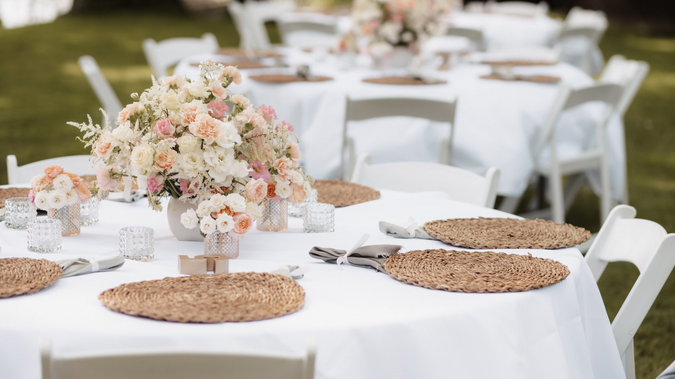 Runde Tische mit weißen Tüchern, floralen Tafelaufsätzen und geflochtenen Tischsets werden in Berlin für eine Veranstaltung im Freien aufgestellt - perfekt für Paare, die mit einem Hochzeitsplaner oder einer Freien Traurednerin zusammenarbeiten.