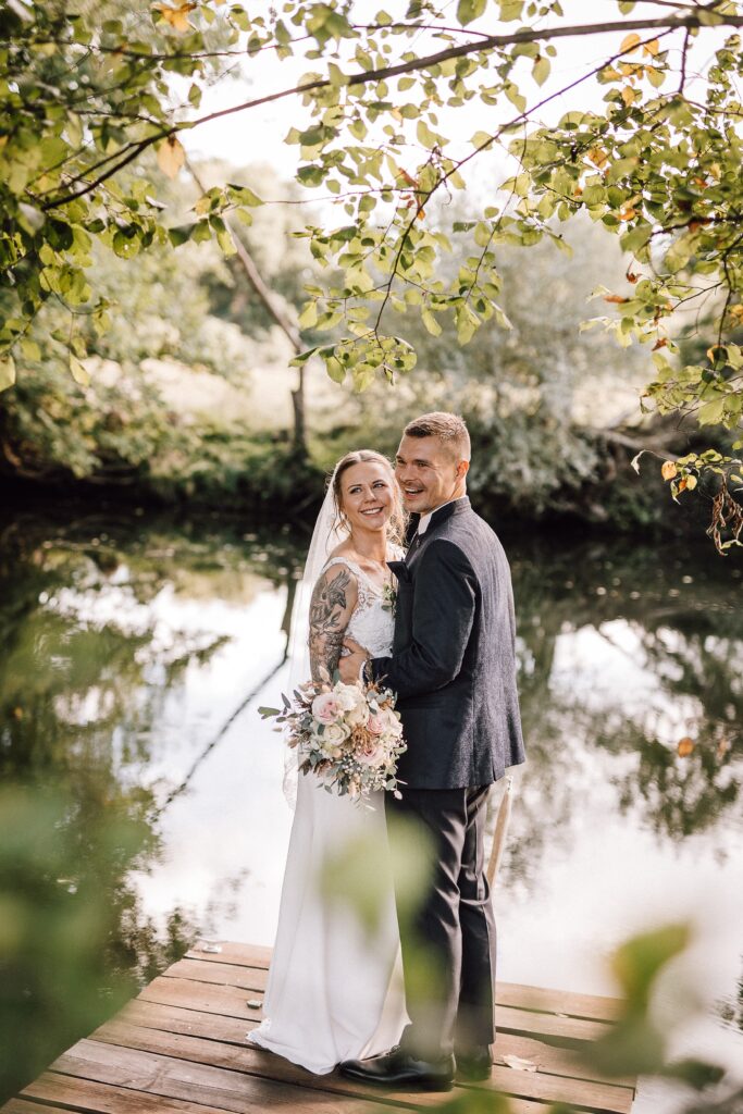 Braut und Bräutigam lächelnd und umarmend auf einem hölzernen Steg an einem ruhigen, von Bäumen gesäumten Teich nach ihrer innigen Zeremonie mit einer Freien Rednerin Berlin.