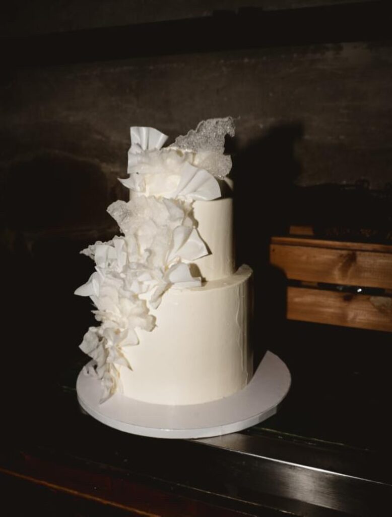 Eine zweistöckige weiße Hochzeitstorte, die auf einer Seite mit großen, zarten weißen Zuckerblumen verziert ist.