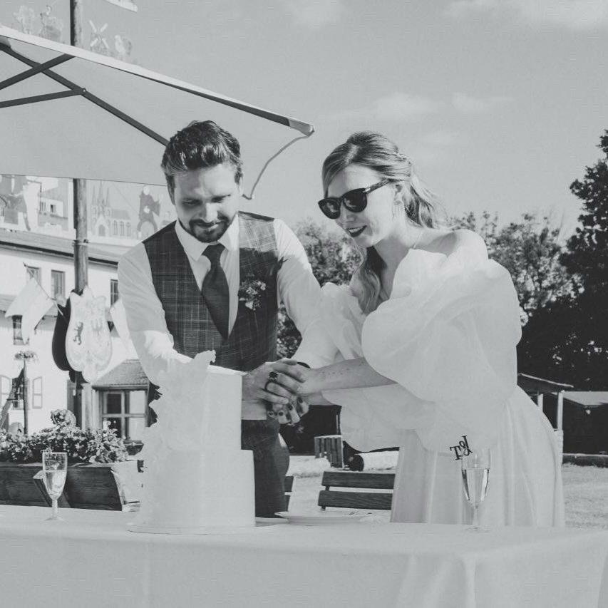 Ein lächelndes Brautpaar schneidet seine Hochzeitstorte im Freien unter einem großen Regenschirm an und feiert mit eleganten Hochzeitstorten an einem schönen Tag.