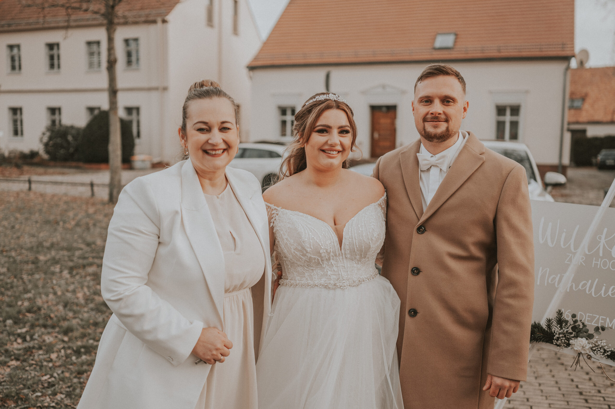 Drei Personen lächeln im Freien auf einer Freien Trauung in Berlin, wobei eine Braut in einem weißen Kleid zwischen zwei anderen Personen in hellen Mänteln steht.